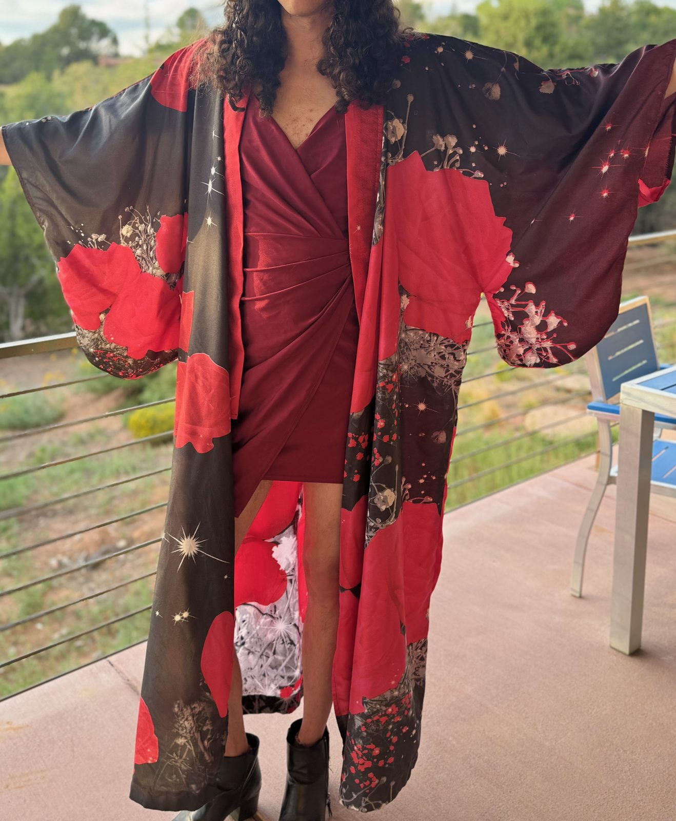 A person on a porch wearing a long dark robe printed with large red poppies, arms outstretched.